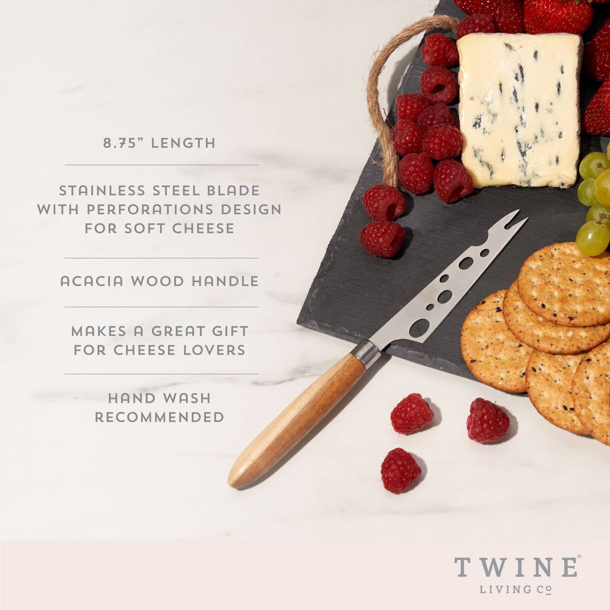 Image of a black slate charcuterie tray with fresh raspberries, soft cheese, green grapes, crackers and a soft cheese knife resting on top. The soft cheese knife has ovals cut out of the blade creating a swiss cheese look and light brown, acacia wood handle.