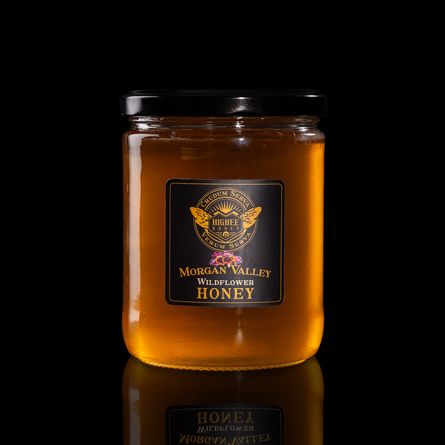 Honey - Morgan Valley Raw, Unfiltered Wildflower Honey Honey  in a 20 oz. glass jar with a black lid on a black background with a fading reflection on a glossy surface. The label is black and gold with Rocky Mountain and Wasatch Range wildflower floral elements behind the seal of Higbee Honey with it's logo in front.