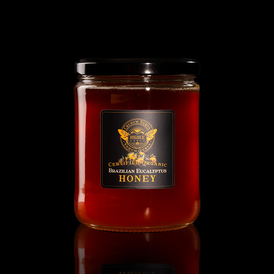 Honey - Brazilian Eucalyptus Organic Honey Raw and Unfiltered 20oz jar Honey with a black lid on a black background with a fading reflection on a glossy surface. The label is black and gold with Brazilian Eucalyptus flower floral elements behind the seal of Higbee Honey with it's logo in front.