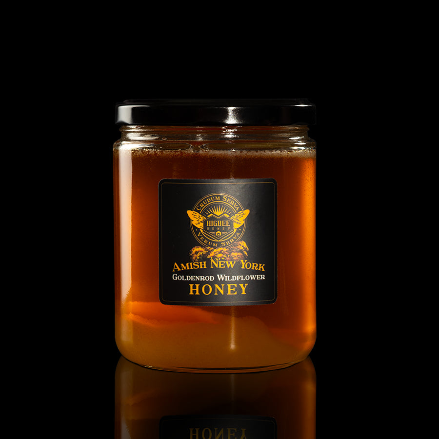 Honey - Amish New York Goldenrod Wildflower Honey in a 20z. Jar with a black lid and black and gold label with goldenrod floral elements in front of a seal of Higbee Honey that includes the Higbee Honey Logo. Jar is on a glossy surface with reflection of the jar and everything is on a black background.
