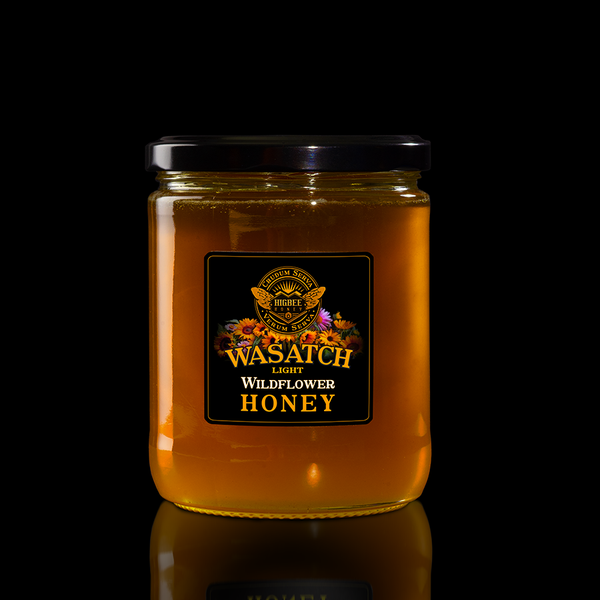Honey - Wasatch Local Wildflower Honey - Light Honey - Wasatch Wildflower Light honey in a glass jar with a black lid on a black background with a fading reflection on a glossy surface. The label is black and gold with Rocky Mountain and Wasatch range wildflower floral elements behind the seal of Higbee Honey with its logo in front.