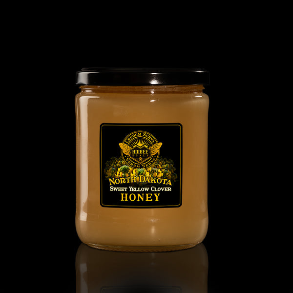 Honey - North Dakota Yellow Sweet Clover Honey - Creamed honey in a glass jar with a black lid on a black background with a fading reflection on a glossy surface. The label is black and gold with clover floral elements behind the seal of Higbee Honey with it's logo in front.