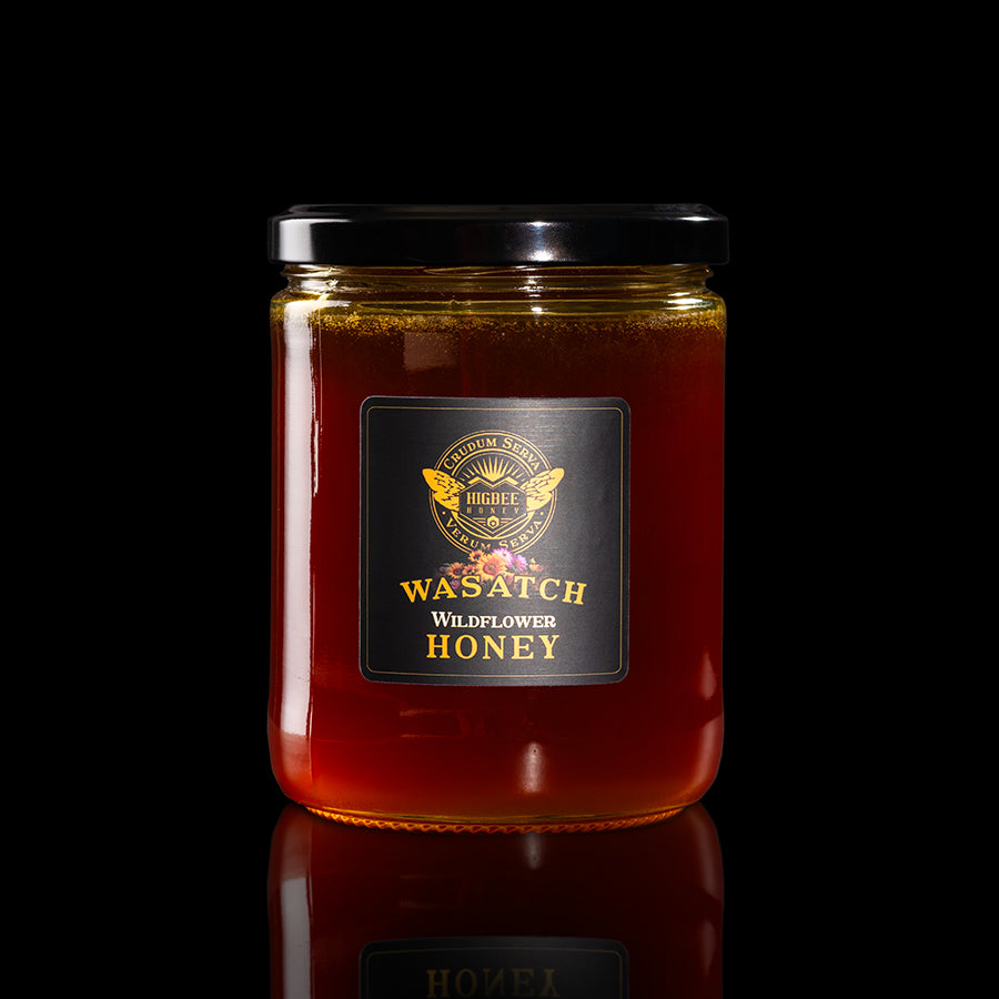 Honey - Wasatch Wildflower Dark honey in a glass jar with a black lid on a black background with a fading reflection on a glossy surface. The label is black and gold with Rocky mountain and wasatch mountain range wildflower floral elements behind the seal of Higbee Honey with it's logo in front.