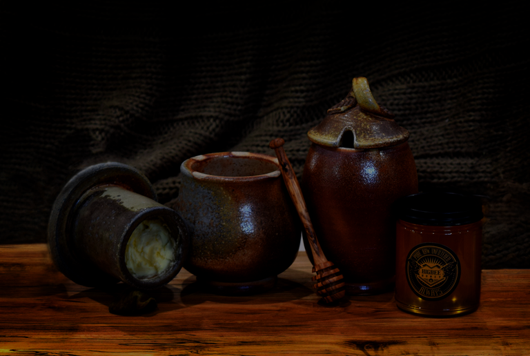 Butter rock with its lid on its side showing butter inside next to a taller honey crock and to its right, a small jar of Morgan Valley Local Raw uniltered wildflower honey. All are sitting on a spalted pecan charcuterie board with a crocheted gray backdrop drapery.